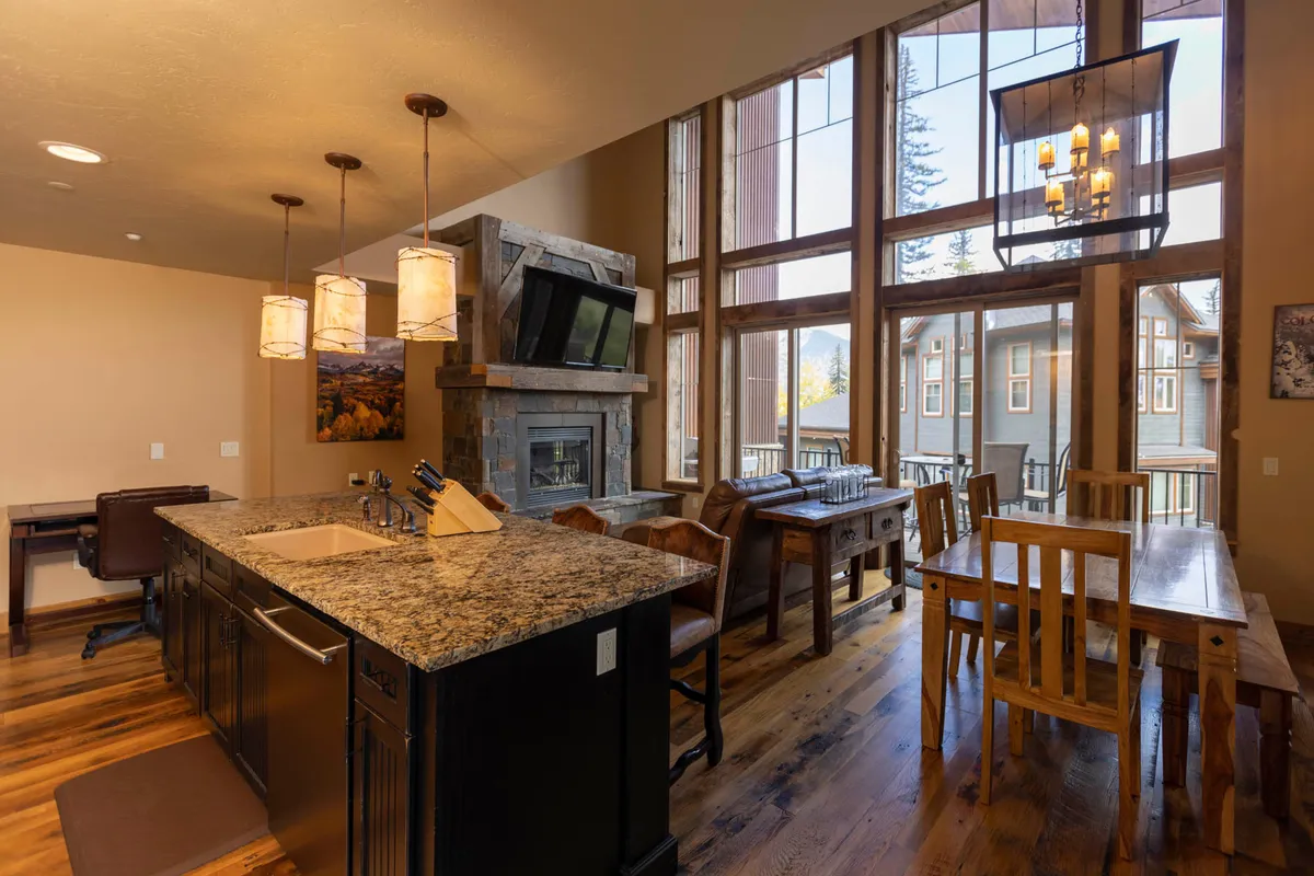 The image showcases a warm, inviting interior featuring a spacious granite kitchen island illuminated by elegant pendant lights, a cozy stone fireplace with a mounted TV, large floor-to-ceiling windows framing breathtaking mountain views, and a wooden dining table perfect for gatherings. The space exudes comfort and connection, with direct access to the slopes visible just beyond the windows.