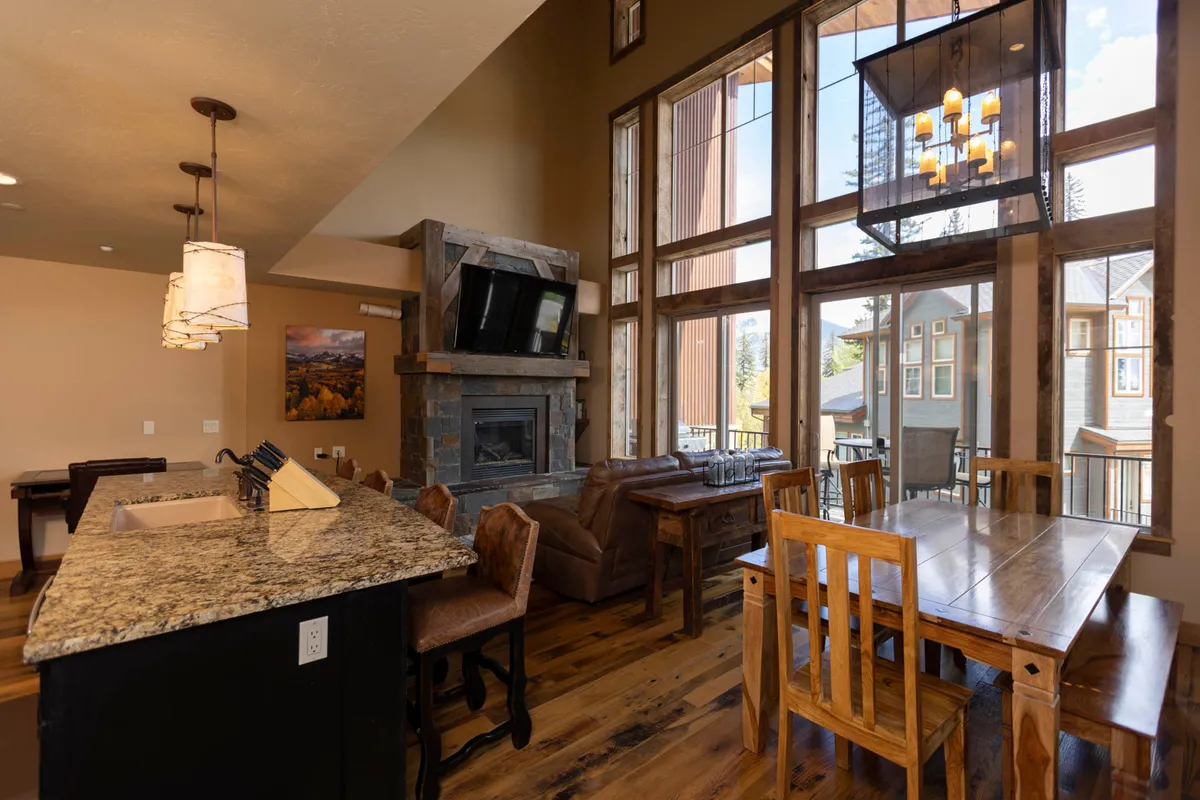 A welcoming open-concept living area featuring a large stone fireplace with a mounted TV, floor-to-ceiling windows providing panoramic views of the mountains, a granite kitchen island with seating, and a wooden dining table with chairs. The space exudes warmth with its plush furnishings and natural light, and there is a direct visual connection to the outdoor deck.