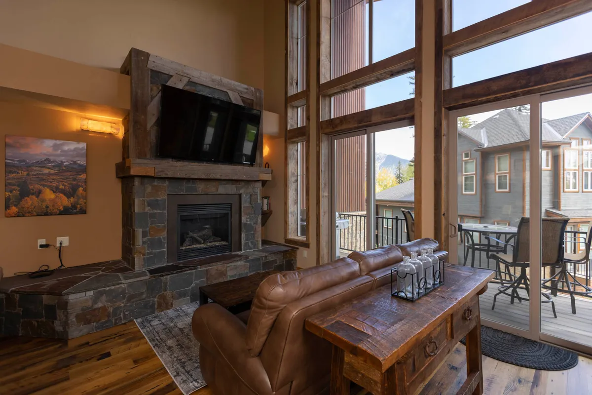 This interior image shows a cozy living area with a stone fireplace, leather sofa, large windows framing mountain views, and a wooden deck with outdoor seating visible just outside. The scene captures the transition from indoor to outdoor living and highlights both relaxation and scenic enjoyment.