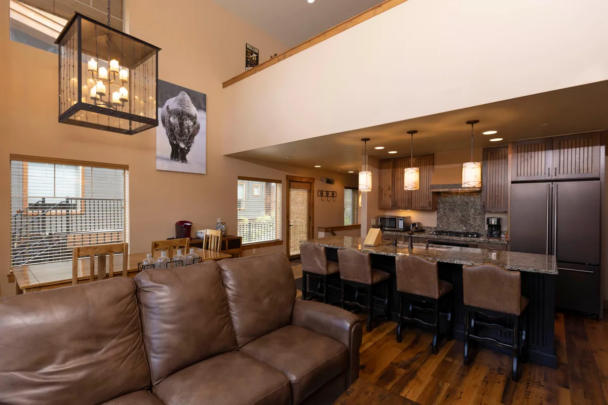 An open living space with a large leather sofa, a kitchen island with bar stools, high vaulted ceilings with a chandelier, and bison artwork on the wall. The scene exudes warmth and comfort—ideal for relaxing, socializing, or utilizing the sleeper sofas for extra guests.