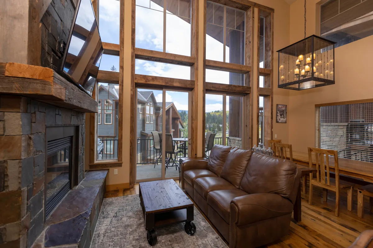 A tranquil living room with large floor-to-ceiling windows framing stunning mountain vistas, a stone fireplace with a mounted TV, a comfortable leather sofa, and a wood dining table under a glowing chandelier. The outdoor balcony is visible, reinforcing the indoor-outdoor connection.