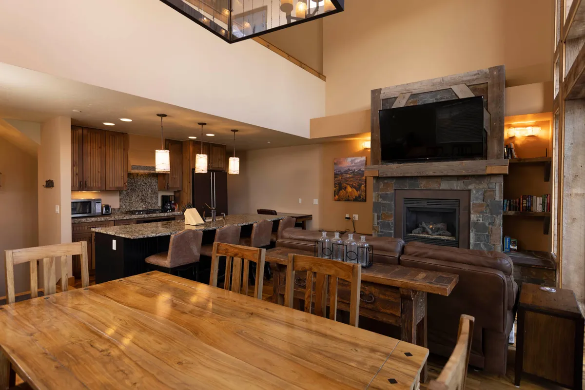 This inviting open-concept space includes a large wooden dining table, a kitchen island with pendant lighting, a stone fireplace with a mounted TV, and a brown leather sofa. The dining area is adjacent to the kitchen, seamlessly connecting meal prep and group gatherings.