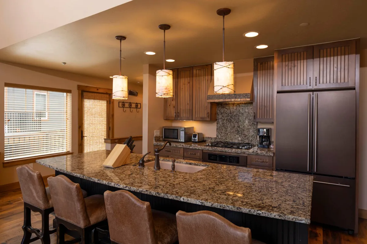 This image features a large granite kitchen island with an integrated sink and seating, a stainless steel refrigerator, and wood cabinetry with vertical grooves. Three pendant lights hang above the island, creating a cozy and functional atmosphere. The space is clearly designed for both meal preparation and casual gathering, with enough room for multiple people to participate in cooking or enjoy snacks and drinks together.