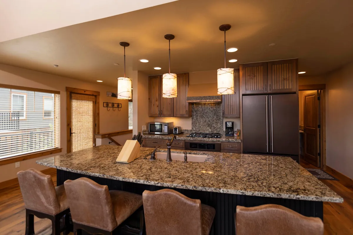 A wide interior shot displays a large granite kitchen island with seating, wood cabinetry, stone backsplash, and modern appliances including a refrigerator and stove. Three pendant lights illuminate the area, and the layout invites group cooking as well as casual dining or conversation.