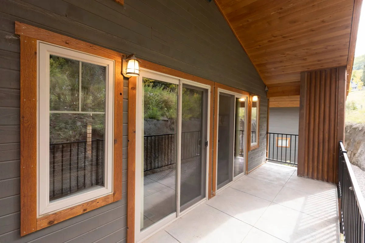 An outdoor deck area with sliding glass doors leading directly into the living space, wood-framed windows, warm wall lamps, and a wood-paneled ceiling. The scene is set for enjoying mountain views, fresh air, and seamless indoor-outdoor living.
