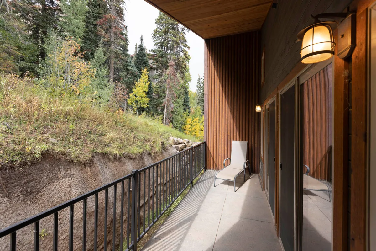 This exterior shot features a narrow balcony with a metal railing, a single lounge chair, wood-paneled walls with warm lighting, and a natural hillside with trees and greenery. The scene evokes a connection to the outdoors and the changing mountain seasons, providing a peaceful setting for relaxation after a day of adventure.