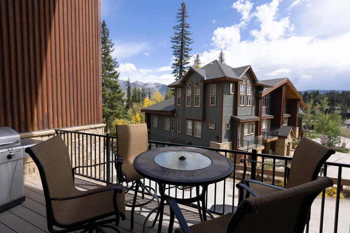 An outdoor patio with a round table and four chairs sits on a balcony overlooking neighboring townhomes and pine trees, with mountains in the distance. A modern barbecue grill hints at family meals and relaxation after outdoor adventures. The scene feels peaceful yet adventurous, with direct access to the outdoors.
