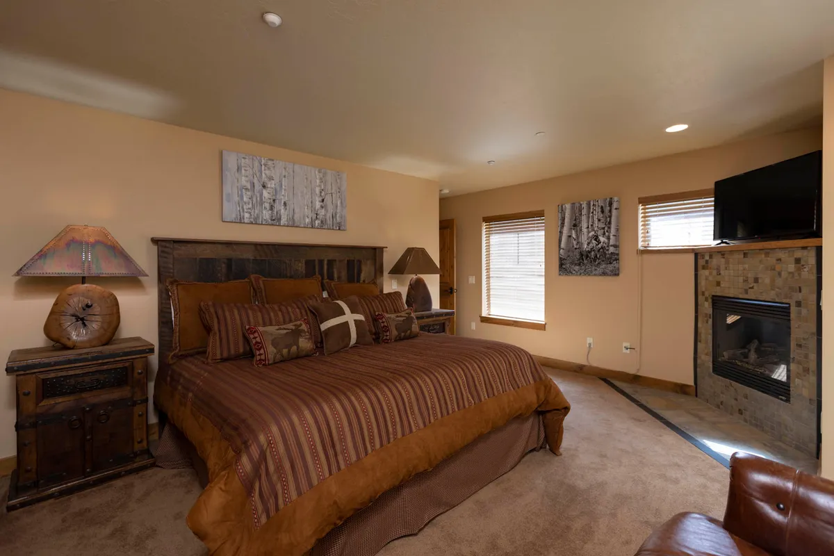 A spacious bedroom featuring a large king-sized bed with a rustic wooden headboard and patterned bedding. The room is enhanced by a cozy corner fireplace with stone tile surround, side tables with decorative lamps, and art depicting birch trees, all bathed in natural light for a peaceful ambiance.