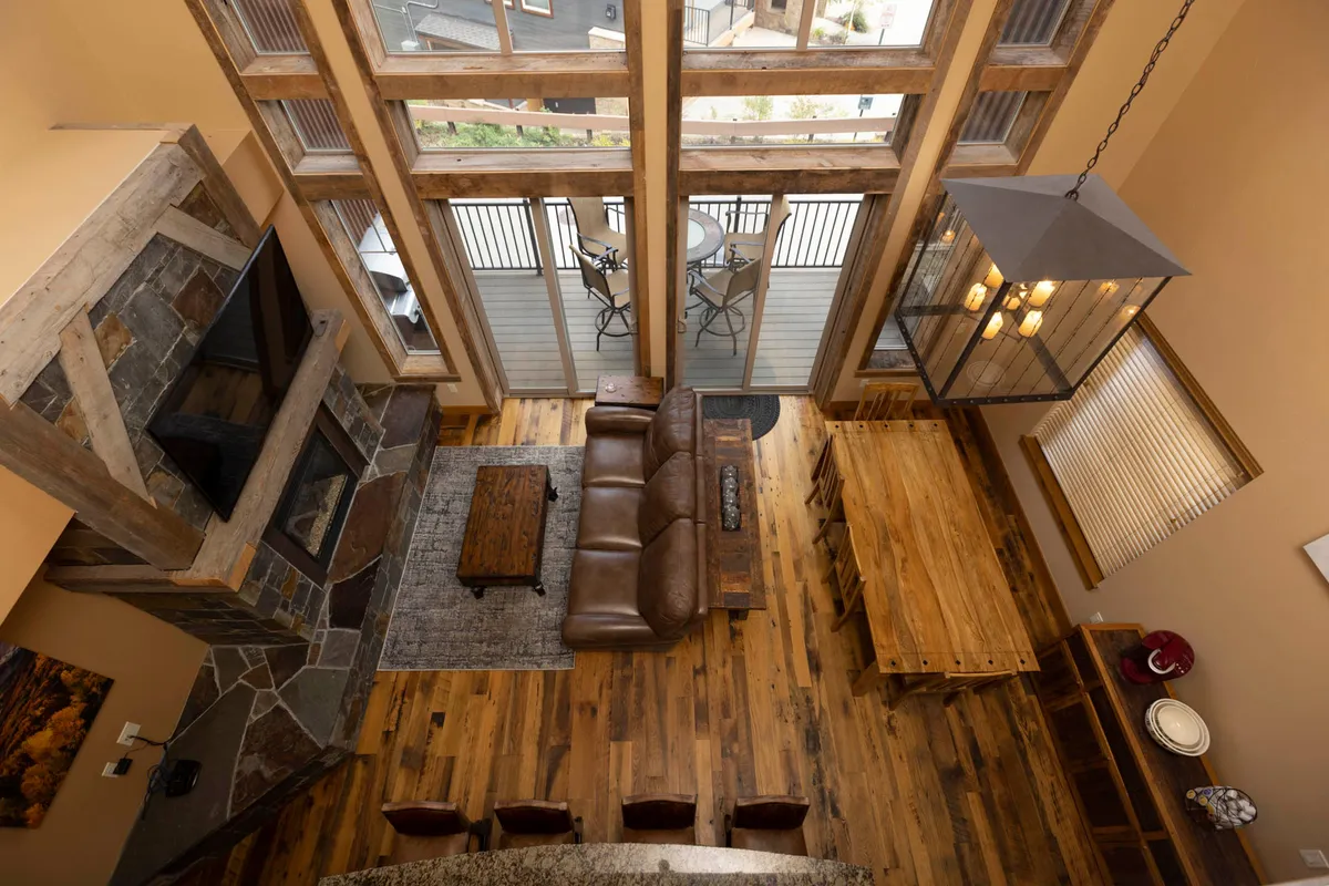A cozy living space featuring a large stone fireplace with a wooden mantel, a leather sofa facing the fireplace and TV, a wooden dining table with chairs, and floor-to-ceiling windows leading to a balcony overlooking the outdoors. The room is spacious and designed for groups to gather comfortably.