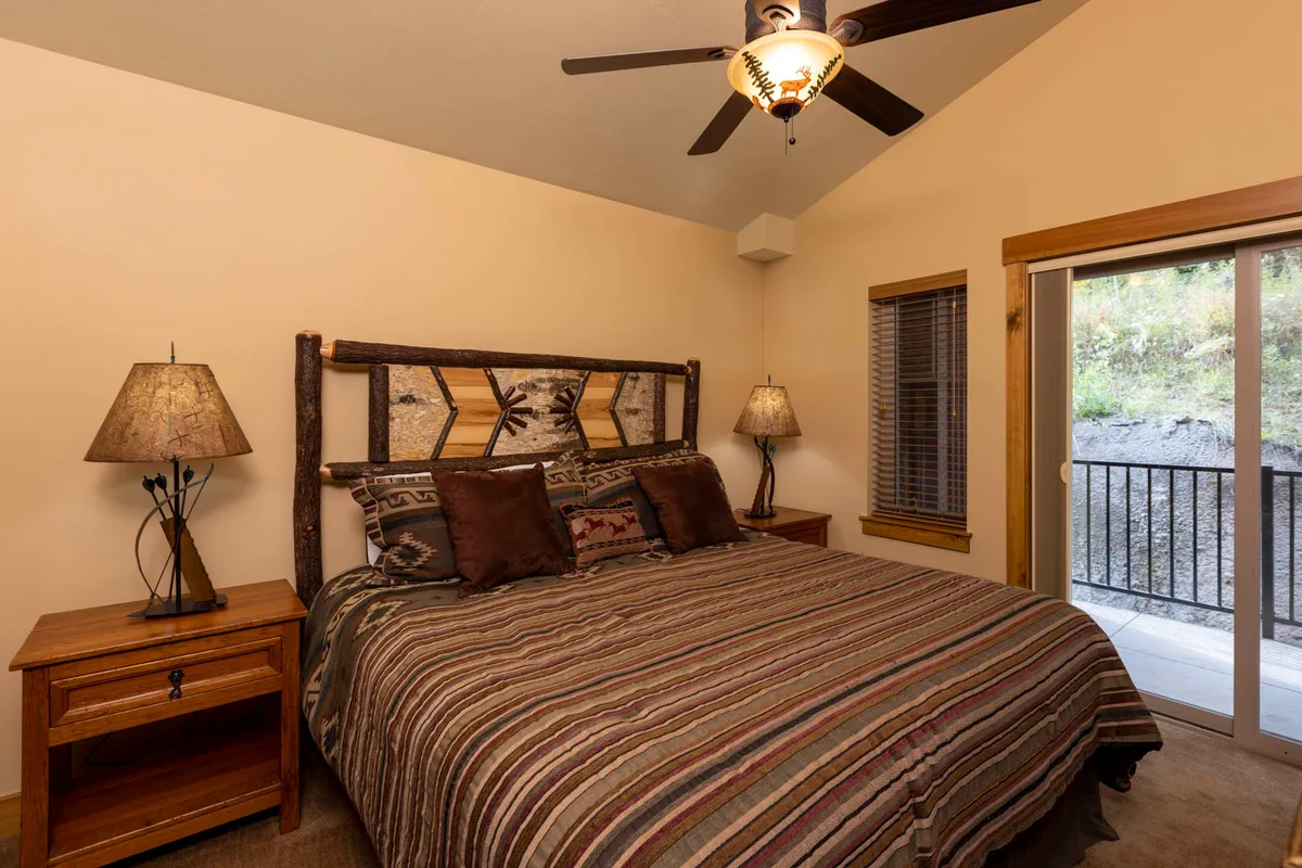 A bedroom with a large bed, rustic wooden headboard, striped bedding, and matching nightstands. The room includes a sliding glass door that opens to a balcony, letting in natural light and offering a glimpse of the outdoors—ideal for morning relaxation or nighttime unwinding.