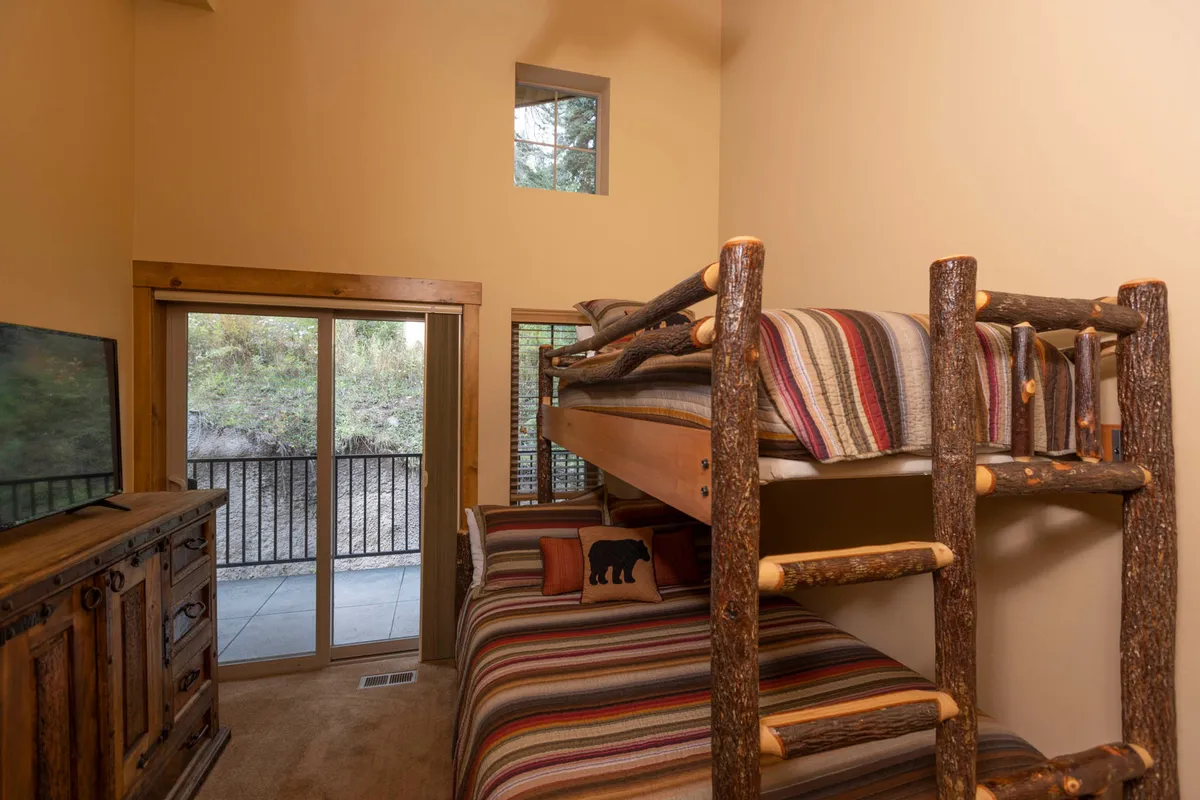 A cozy bedroom with a rustic log bunk bed dressed in striped bedding, a wooden dresser with TV, and a sliding glass door leading to a balcony. The space is designed for multiple guests, making it perfect for kids or friends who want to bunk together.