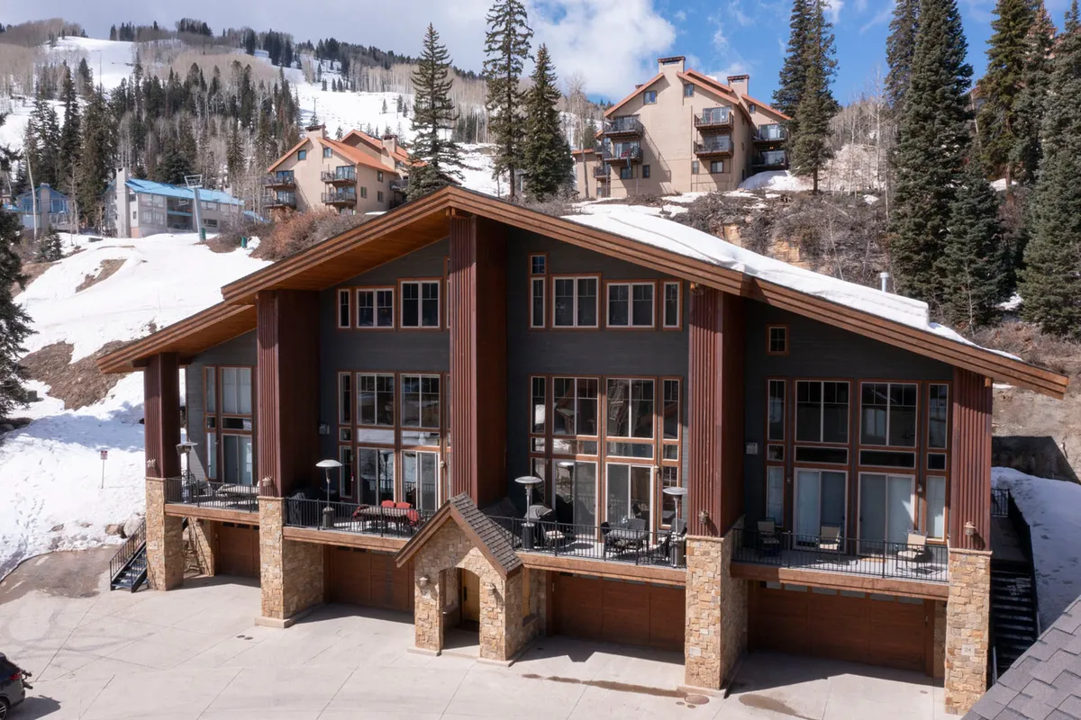 A wide view of a large townhome with peaked roofs and multiple balconies, surrounded by snowy pine trees and directly neighboring ski slopes. The property’s stone and wood facade blends into the natural alpine environment, and the snow-covered landscape conveys a cozy mountain escape.