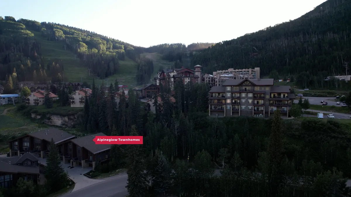 A wide shot of the Alpineglow Townhomes complex nestled among lush evergreen trees, with the ski slopes and mountain peaks visible in the background. The image radiates a serene, inviting mood, and highlights the townhome's location directly adjacent to the slopes.