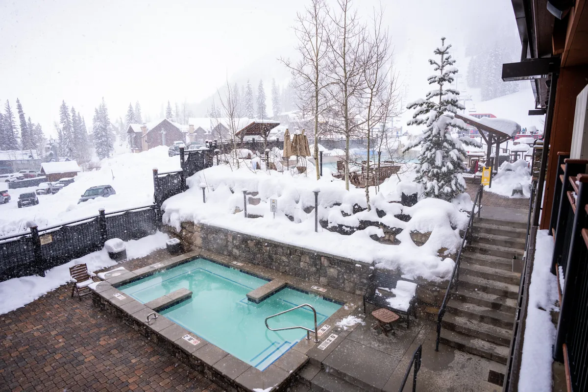 A heated outdoor pool is surrounded by snow-covered landscaping, trees, and stone retaining walls. In the background, mountain resort buildings accentuate the alpine setting. People are seen soaking in the warm pool, enjoying the contrast between the inviting water and the snowy environment.
