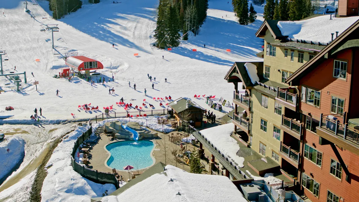 This dynamic exterior photo features skiers on snowy slopes, an outdoor heated pool with a slide, a multi-story lodge with balconies, and a ski lift station. The energetic scene captures both winter sports action and post-adventure relaxation, with guests enjoying the pool and resort amenities.