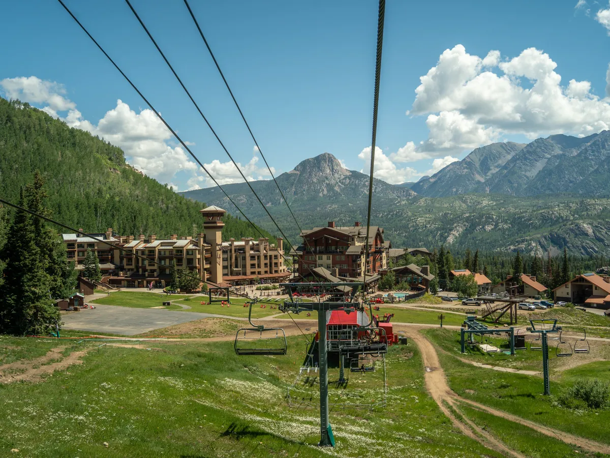 This exterior image captures ski lift chairs gliding above grassy slopes, mountain lodge buildings nestled at the base, and a dramatic mountain range in the background. The setting conveys the energetic yet peaceful atmosphere of a mountain resort, hinting at both winter and summer activities, and the direct connection between lodging and outdoor adventure.