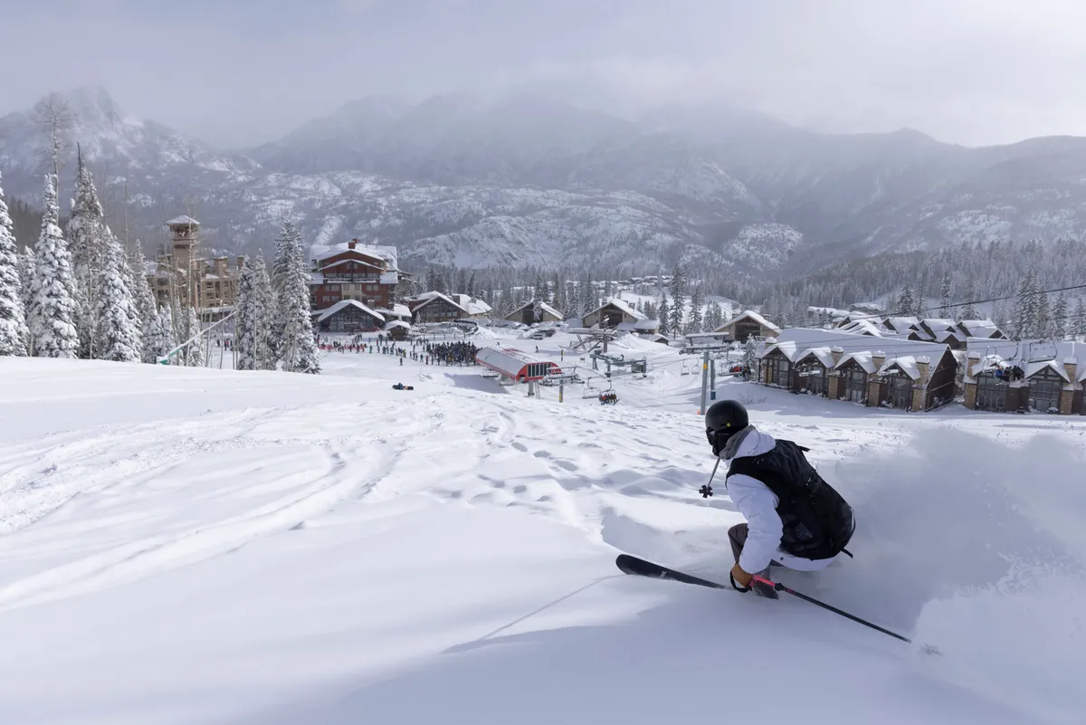 A skier carves fresh powder on a bunny slope directly in front of a row of snow-covered townhome buildings. The ski lift and gentle beginner slopes are visible, with mountain peaks creating a stunning backdrop. The scene radiates winter fun and family-friendly adventure, with easy access between the townhomes and the slopes.